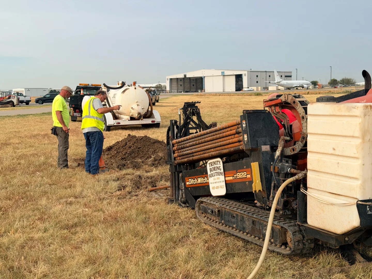 Trinity Boring Solutions crew running a 2 inch conduit directional bore with airport hangars and a parked jet in the background in Oklahoma City
