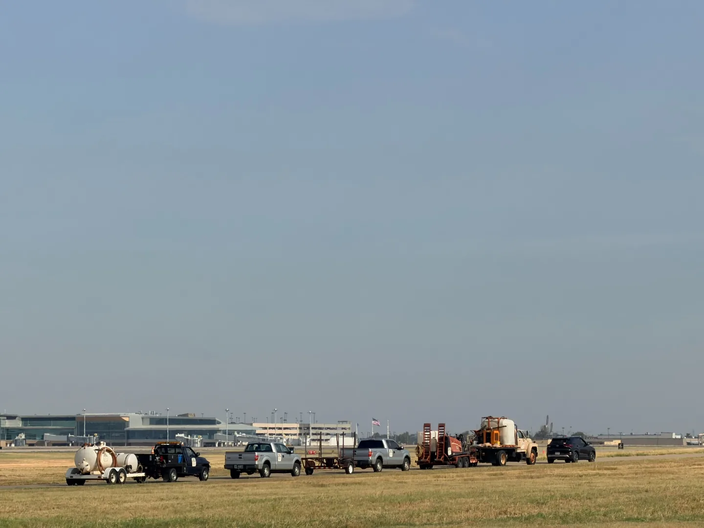 Trinity Boring Solutions drill rig, hydrovac trailer and support trucks staged along an airport service road with the Will Rogers World Airport terminal in the background